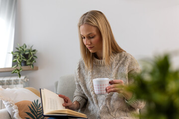 Woman Reading Book and Drinking Coffee on Sofa, Cozy Morning Routine Concept. Blonde female in warm sweater holding mug and enjoying literature in modern home living room environment.