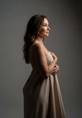Fototapeta premium Graceful profile portrait of brunette woman in strapless beige drape, minimalist fine art studio lighting on gray background