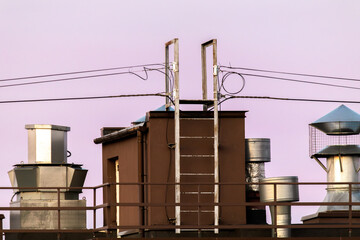Industrial ventilation units and a rooftop access ladder sit beneath overhead wires, suggesting maintenance and technical systems that keep a building running. The minimalist sky and geometric lines c