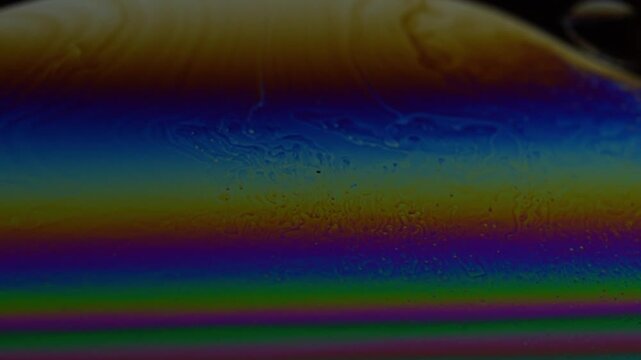 Macro video of a soap bubble surface showing dancing iridescent colors and smooth fluid motion. Abstract liquid patterns shift organically across a dark background, creating  ethereal, calming visual