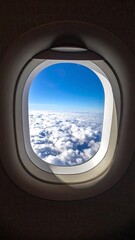 Plane window shows bright blue sky over fluffy white clouds from a high vantage point