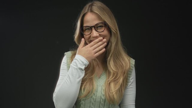 Woman with long blonde hair and round glasses covering her mouth laughing in studio wearing green vest and white long sleeves; amusement surprise joy happiness.