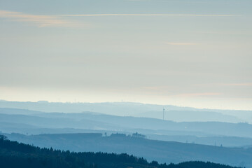 Fototapeta premium Atmospheric landscape of rolling hills gradually fading into mist under a pale sky. Soft morning light creates depth and calm mood, with wind turbines visible on the distant horizon.