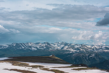 Obraz premium Snowfields and thawed ground on hill top. Sharp rocks on precipice edge above forest valley with view to big snowy mountain range far away under cloudy sky. Sunlight and shadows in high mountains.