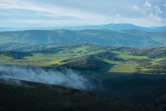 Scenic aerial top view from above through low cloud on pass to green hilly vastness and layered mountain silhouettes far away under dramatic cloudy sky. Sunlight and shadows of clouds on forest hills.