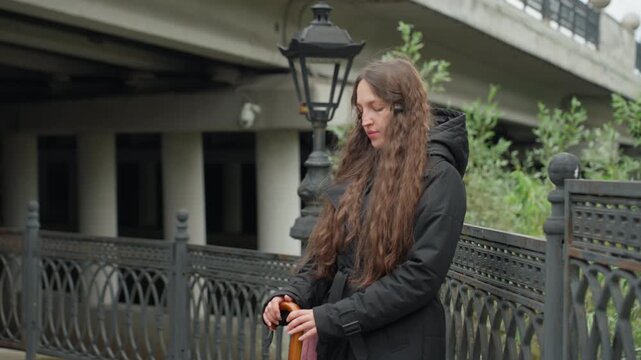 full frame white woman by railing, standing under overpass with long brown hair and black coat, holding small object near iron fence and vintage lamp, urban commuter pause with contemplative gaze,