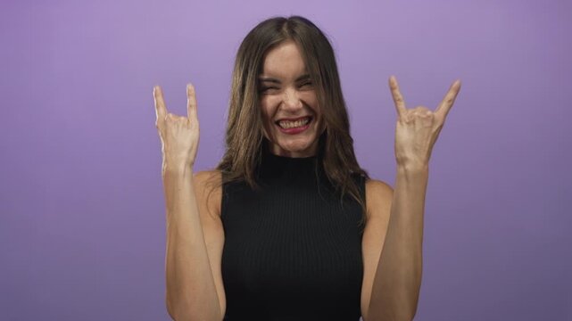 Woman smiles and shows rock horns gesture with both hands and bare arms in purple studio; rock fun music excitement.