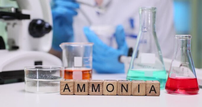 Wooden cubes spell word Ammonia on science lab table near glassware. Scientist in gloves works with flasks testing chemical reaction using microscope