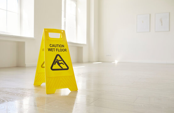 Caution wet floor sign warning of slippery danger in empty office room. Yellow plastic safety and prevention signal of professional janitor standing indoors. Cleaning service, precaution concept