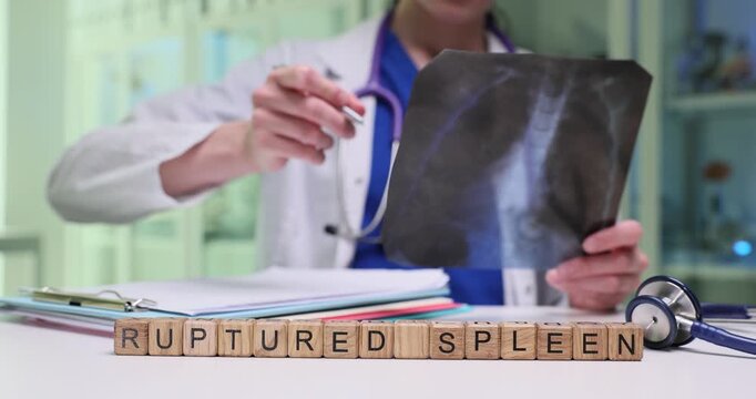 Wooden cubes spell phrase Ruptured Spleen on medical desk. Doctor studies chest X-ray holding film while writing report about abdominal trauma case