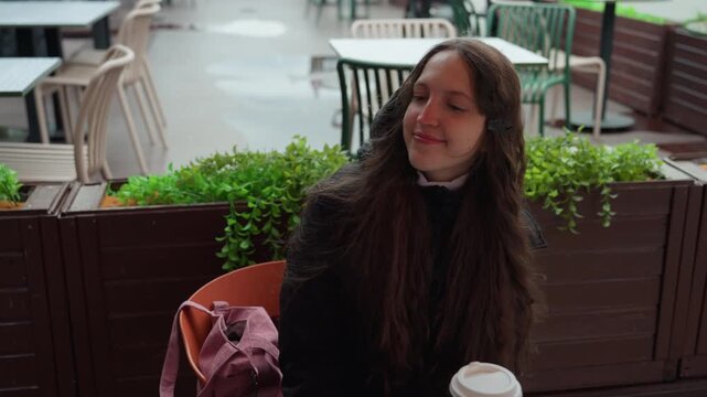 caucasian woman walking rainy cafe terrace, settling at table to read journal, long brunette hair under hood, parka damp from drizzle, hands opening book, takeaway coffee on table, commuter pause