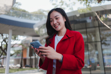 Fototapeta premium Smiling young Asian woman networking on a smartphone in a modern urban area, representing communication and social media