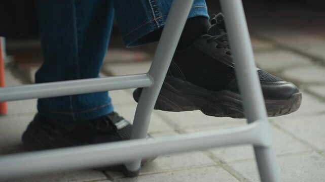 black sneaker under metal stool on pavement, closeup of leg in blue jeans resting on stool rung, outdoor cafe or sidewalk setting, casual commuter pausing midwalk, textured rubber sole and scuffed