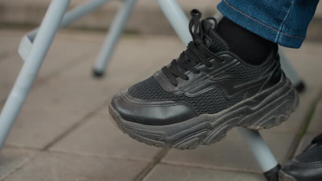 black sneakers on tiled pavement, commuter foot tapping under metal chair, denim cuff visible, laced trainer with worn sole and scuff marks, casual waiting scene suggesting walking break
