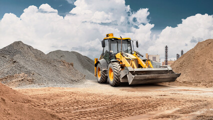 Obraz premium Loader transports sand using its storage bucket at a construction site with large piles of materials and heavy machinery visible in the background