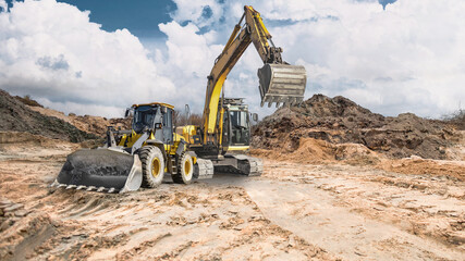 Obraz premium A loader and a crawler excavator work at a construction site. In the background, earthen embankments rise under a partly cloudy sky