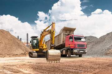 Obraz premium An excavator moves sand from a mound into a truck at a busy construction site. Heavy equipment performs earthmoving and bulk material handling under a clear blue sky