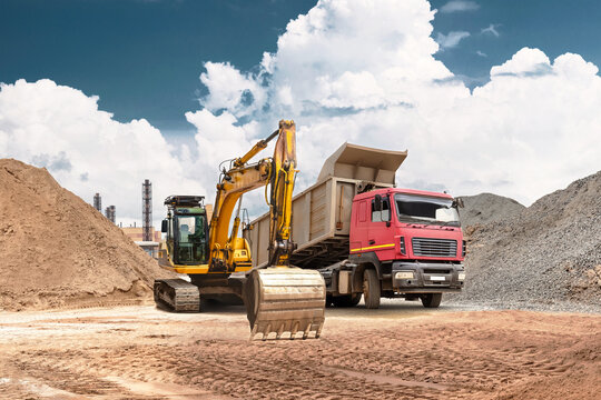 An excavator moves sand from a mound into a truck at a busy construction site. Heavy equipment performs earthmoving and bulk material handling under a clear blue sky