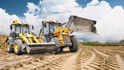 Two construction loaders are ready to go to work at a construction site, and a large pile of sand is visible against the cloudy sky. The work continues