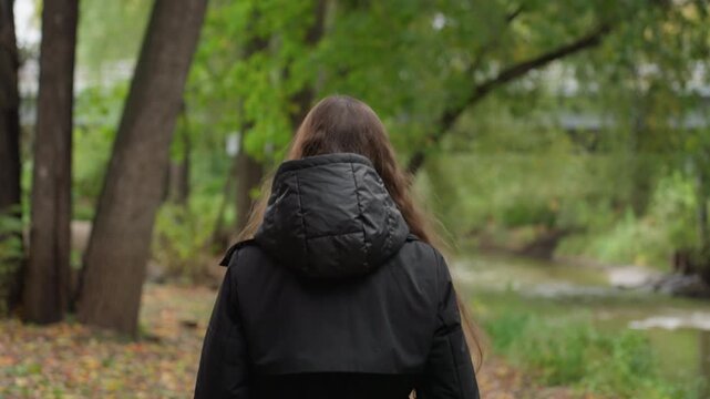 caucasian woman turning walking away from camera backview in hooded jacket strolling along creek path hands in pockets slow steps autumn foliage urban bridge ahead quiet exploration vibe ideal