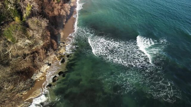 Aerial drone view of sea waves moving along a rocky coastline with a narrow sandy shore. Clear green-blue water reveals underwater rocks as white foam patterns form near the shore. 