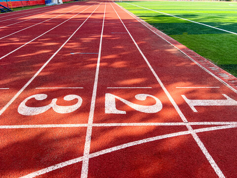 Red rubber running track lanes with start numbers 1, 2, 3 and adjacent green artificial grass field. Authentic outdoor stadium surface, ideal for racing, sports training, and competition backgrounds.