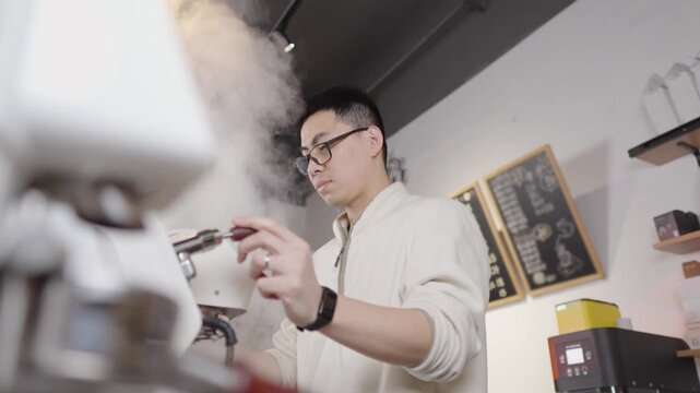 Skilled asian barista wearing glasses using a professional espresso machine to steam and froth milk for cappuccino or latte in a modern coffee shop, with steam rising in slow motion