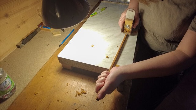 Hands shaping a wooden bow stave with a spokeshave on a lit workbench showing wood shavings
