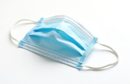 Light blue disposable face mask with ear loops rests on clean white surface. Protective item shields against viruses, dust, air pollution. Essential for health, safety measures. People use for