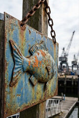 Fototapeta premium A weathered blue metal fish sign with peeling paint hangs from a rusty chain on a wooden post at a harbor.