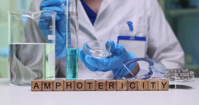 Wooden cubes spell word Amphotericity on desk near glassware. Scientist uses pipette and liquids during chemistry experiment in bright laboratory