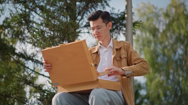 young white man opens wooden sketchbox, under pine trees with sunlight, assembles portable easel, arranges brushes charcoal and paper, sits on stool and studies landscape, begins careful pencil