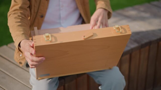 man opening wooden sketch box in urban park, preparing portable painting setup, empty interior with hinged compartments, casual jacket and jeans, warm sunlight highlights natural wood texture, hands