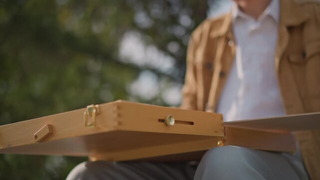artist opening wooden sketch box outdoors, sits on bench in sunlit park, unfolds pochade panel, inspects board and brass latches, hands in closeup, wearing casual jacket, atmosphere of focus