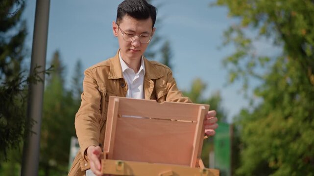 young male artist outdoors setting easel and canvas, wooden sketch box opened on lap, hands arranging brushes and bottle, concentrated expression under warm sunlight, park trees and street pole