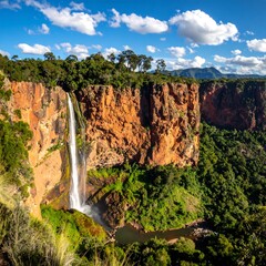 A majestic waterfall cascades down a rugged, red-hued cliff face, surrounded by lush green foliage under a bright, cloud-filled sky