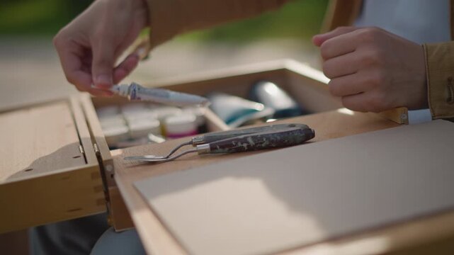 closeup hands closing wooden art box on lap, canvas placed inside and smoothed, brass latch turned to secure supplies, quiet park backdrop with soft sunlight, ritual end of creative outdoor painting