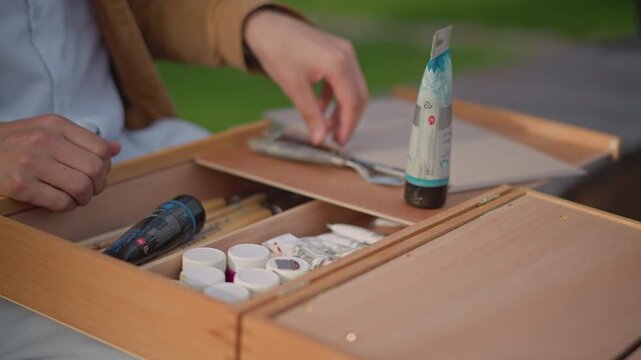 white artist arranges wooden paintbox on bench, selecting tubes, palette knife and brushes, organizing compartments, closing latch, soft natural light, street art sketchbook hint, calm reflective