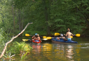 Back view of unrecognizable people in colorful kayaks paddling down a Krutynia river through a dense green forest. Summer group kayaking trip.