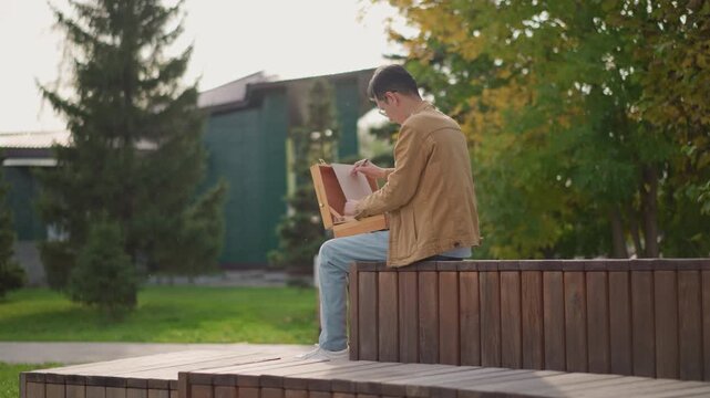 young man drawing architecture outdoors on wooden bench in park, midshot showing campus buildings and pines, wearing brown corduroy jacket and jeans, focused gesture, plein air practice, sunset light,