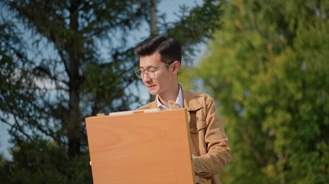 Young artist painting on easel outdoors, concentrated student sketching sunlit landscape in leafy park with wooden sketchbox brown jacket, closeup on hands and brushwork, textured strokes on canvas,