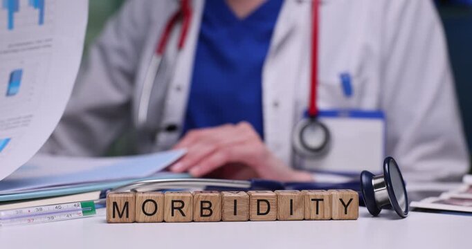 Wooden blocks spell word Morbidity on desk near stethoscope and doctor. Doctor reviews patient chart tracking health outcomes in report papers