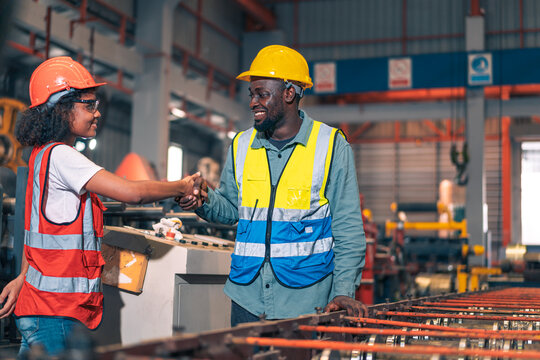 African factory worker man woman shake hand success. Happy male female industrial engineer partner wear safety helmet vest in manufacturing plant. Heavy metal machine background. Diversity teamwork.