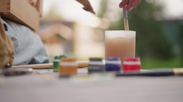 outdoor painting setup with brush dip into water cup, pots of colorful paint lined on wooden table, denim apron and paintbrush visible, soft bokeh backyard and warm sunlight, gentle slow creative mood