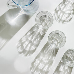 Transparent Water Glasses and water jug with shadows at sunlight on white, pastel color photo minimal style, aesthetic top view shiny cups sparkling at natural light, summer drink, composition