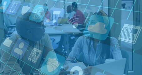 Discussing pair wearing blue shirt and sweater at shared table, using laptop, pen, digital icons