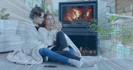 Naklejka premium Cuddling two women in knit sweaters, jeans, under blanket in living room holding mug near fireplace