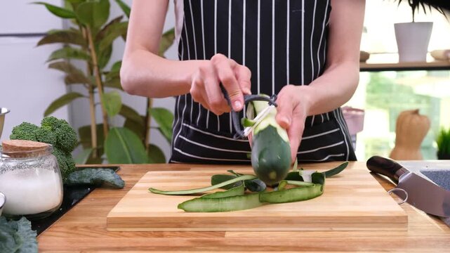 Person peels zucchini on board dropping thin green strips beside peeler. Cook scrapes skin fast turning zucchini to expose fresh surface time-lapse