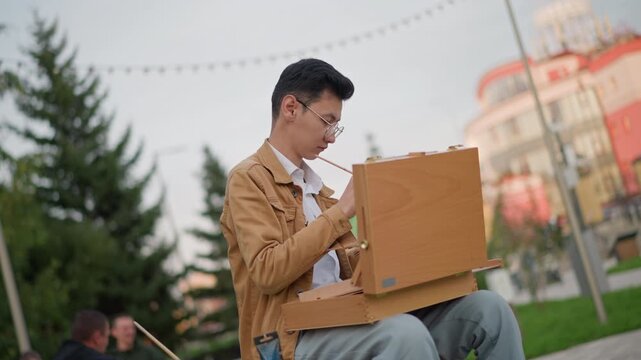 asian illustrator painting in urban park with wooden box easel, focused on composition under string lights, evergreen trees and pastel building in background, people blurred on benches, relaxed