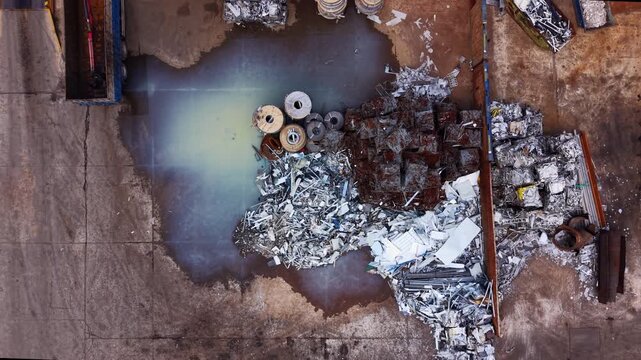 Top-down view of an urban metal scrap yard showing contaminated runoff and toxic puddles formed by industrial waste amid sorted metal stockpiles